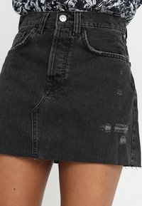 Mini gonna in denim nera a vita alta con tasche frontali, dettagli strappati e orlo grezzo, indossata con un top fantasia.