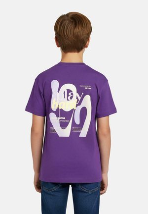 Jongen met kort bruin haar, draagt een paarse T-shirt met witte en gele abstracte tekstprint en een blauwe spijkerbroek, rug naar de kijker, effen achtergrond.