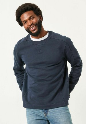 Navyblaue Sweatshirt mit Rundhalsausschnitt, strukturierter Oberfläche, gerippten Bündchen und einer horizontalen Nahtdetails über der Brust. Getragen mit hellblauen Jeans.