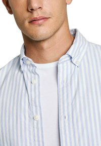 Camisa de botones a rayas en azul claro y blanco con un cuello cerrado y textura suave, llevada sobre una camiseta blanca básica.