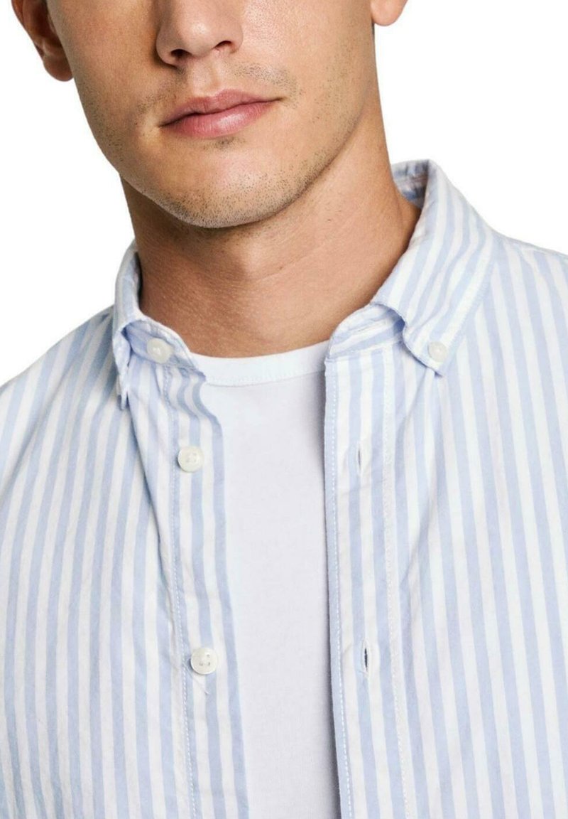 Camisa de botones a rayas en azul claro y blanco con un cuello cerrado y textura suave, llevada sobre una camiseta blanca básica.