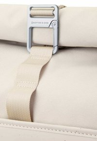 Beige textilní taška s texturovaným povrchem, obsahující stříbrnou kovovou sponu a odpovídající popruh. Na sponě je vygravírováno "KAPTEN & SON."