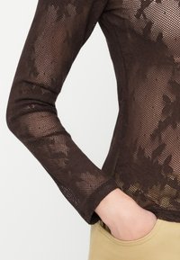 Persona che indossa un top a maniche lunghe in pizzo trasparente marrone scuro con motivi floreali, con la mano appoggiata nella tasca dei pantaloni beige.
