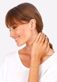 Gold stud earrings with an infinity design, worn on the ear. Simple chain bracelet with an infinity charm. Model has brown hair, wearing a white top.