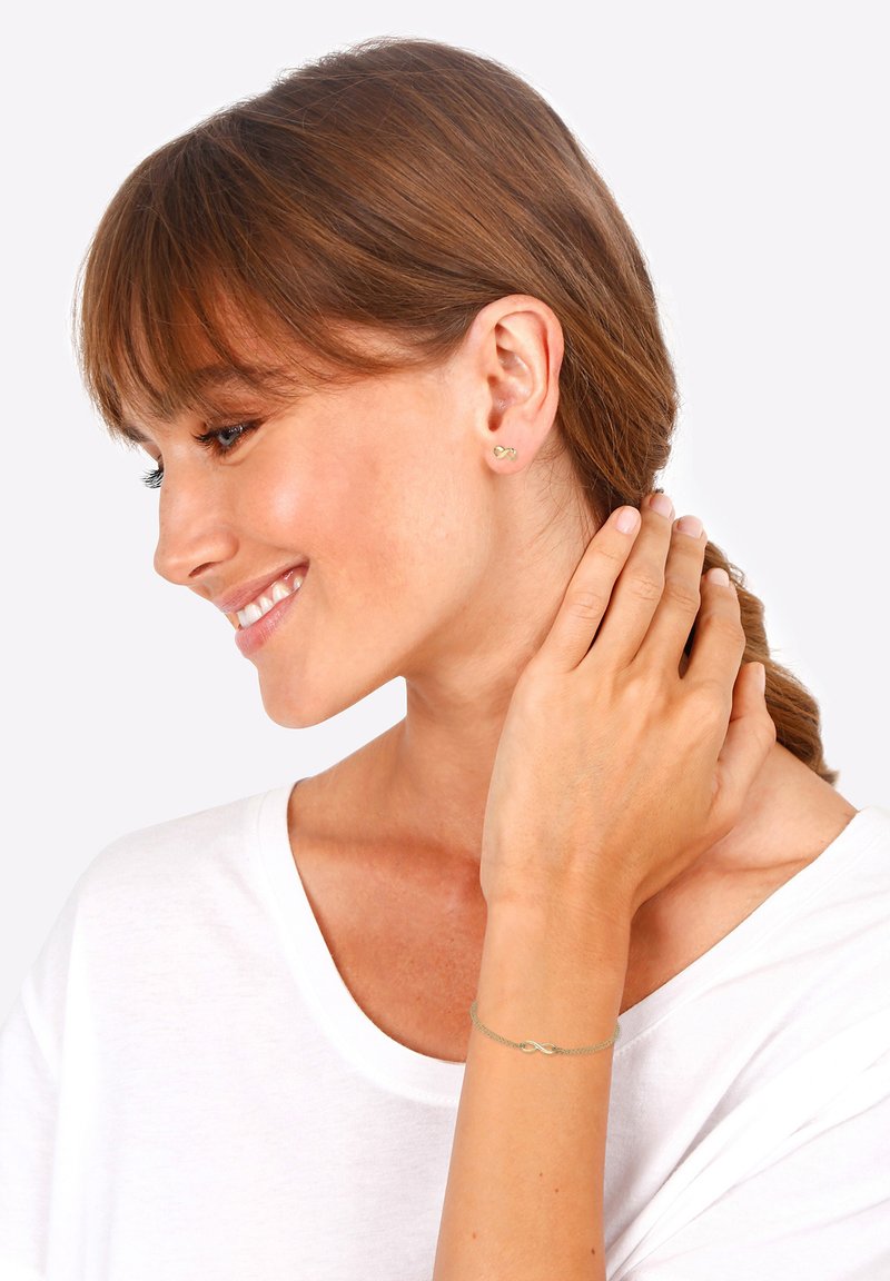 Gold stud earrings with an infinity design, worn on the ear. Simple chain bracelet with an infinity charm. Model has brown hair, wearing a white top.
