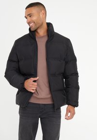 Zwarte pufferjack met ribbelmanchetten en staande kraag, voorzien van een ritssluiting aan de voorkant en een doorgestikt ontwerp. Draagt over een bruine shirt.