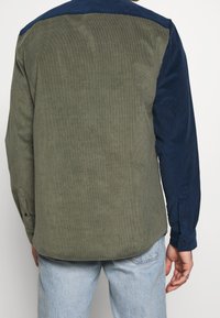 Olivinvihreä ribbatusta sametista valmistettu paita, jossa on laivastonsininen yläosa. Pitkät hihat, joissa nappiratkaisu manseteissa. Rakenteellinen kangas pystysuuntaisella ribbiraidalla.