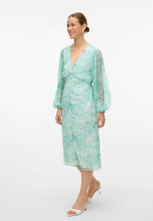 Un vestito verde menta semi-trasparente con scollatura a V, lunghe maniche trasparenti e un motivo floreale, che si estende fino a metà polpaccio.