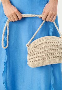 Borsa a mano in crochet beige dalla forma rotonda, con un design a trama aperta e una tracolla intrecciata, tenuta da una mano davanti a uno sfondo di tessuto blu.