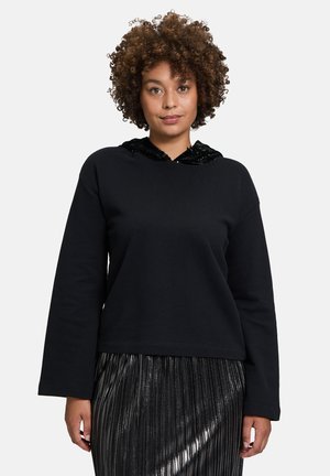 Schwarzer Sweatshirt mit einer Kapuze aus schwarzen Pailletten, langen Ärmeln und einem lockeren Schnitt. Trägt dazu einen silbernen, plissierten Rock.