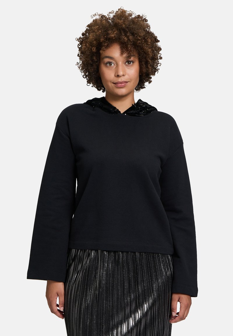 Felpa nera con cappuccio di paillettes nere, maniche lunghe e vestibilità ampia. Indossata con una gonna plissettata argento.