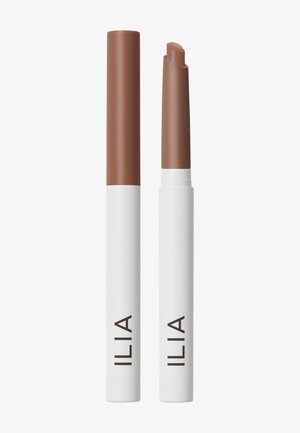 Deux tubes blancs de crayon à lèvres ILIA sont debout, l’un est fermé et l’autre ouvert, révélant un rouge à lèvres marron à l’intérieur.
