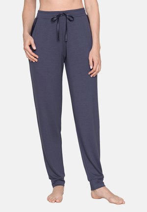 Donna che indossa pantaloni blu scuro con coulisse, in piedi a piedi nudi su sfondo uniforme.