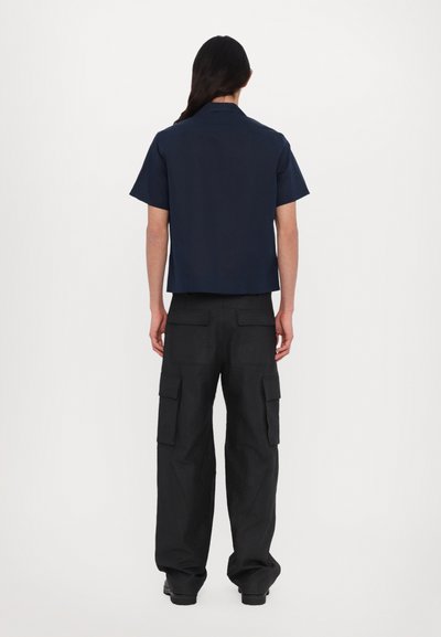 Eine Person steht mit dem Rücken zur Kamera, trägt ein navyblaues kurzärmliges Hemd und schwarze Cargo-Hosen mit mehreren Taschen vor einem schlichten weißen Hintergrund.
