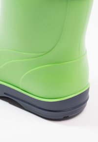 Botte en caoutchouc verte avec une texture lisse, présentant une semelle extérieure bleu foncé, des détails de conception courbés et un haut tige pour une protection contre l'eau.