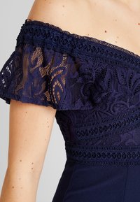 Marinblå klänning med avtagna axlar och intrikata spetsärmar, med blommönster och texturerade band, tillverkad av mjukt tyg.