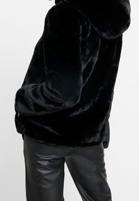 Schwarze Kunstfelljacke mit Kapuze, die über eine weiche Textur und mit Reißverschlüssen versehene Taschen verfügt, kombiniert mit eleganten schwarzen Lederhosen.