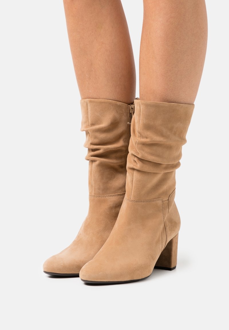 Bottes mi-mollet en daim beige avec tiges effondrées, fermeture éclair latérale, talons carrés, portées sur jambes nues sur un fond clair uni.