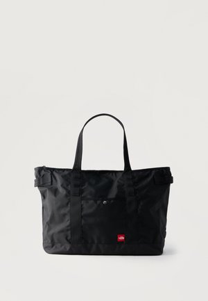 Melna taisnstūra formāta tote soma ar divām rokturiem, priekšējā kabata ar sprādzi un nelikents sarkans logotipa tagiņš apakšējā labajā stūrī.