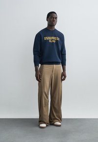 Hombre de pie con una sudadera azul marino con texto amarillo "DSQUARED2 MILANO", pantalones anchos beige y zapatillas blancas. Fondo liso.