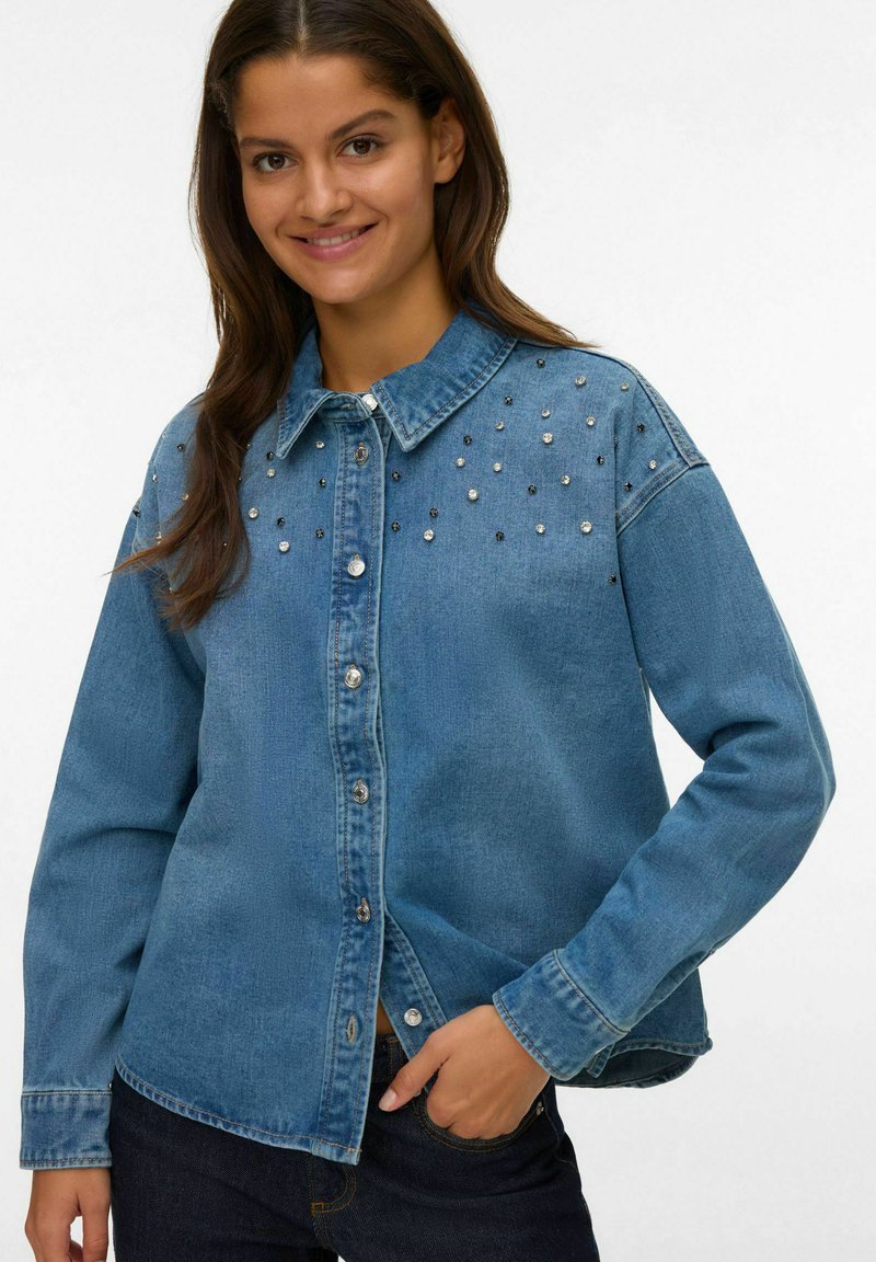 Giacca di jeans in azzurro chiaro con chiusura a bottoni, colletto e impreziosita da borchie argento e nere sulle spalle. Maniche lunghe.