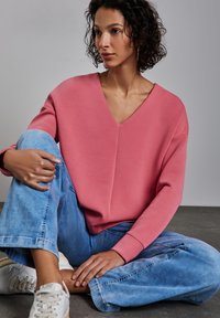 Roze V-hals sweatshirt met verlaagde schouders, gecombineerd met lichtblauwe wijde spijkerbroeken en witte sneakers. Glad materiaal, losse pasvorm.
