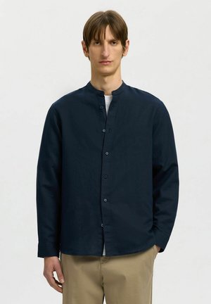 Muž v tmavomodrej košeli s mandarínskym golierom, zapínanej na gombíky, cez biele tričko a béžové nohavice, stojaci na jednoduchom pozadí.