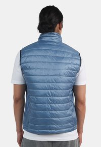 Blå, lätt puffy väst med horisontellt quiltat mönster och hög krage. Har elastiska ärmhål och en rak nederkant. Strukturerad tyg.