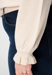 Lichtbeige trui met gestructureerde stof, voorzien van volumineuze, gerimpelde mouwboorden, gecombineerd met donkerblauwe spijkerbroek.