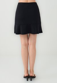 Zwarte, uitlopende mini-jurk met een gladde textuur, voorzien van een aansluitende taille en een gerafelde zoom. Gepaard met zwarte hakken sandalen.