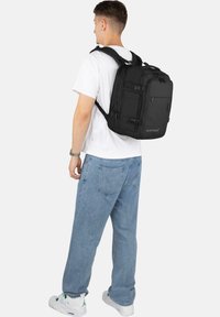 Schwarzer Nylon-Rucksack mit mehreren Fächern, verstellbaren Trägern und einem verstärkten Griff, getragen von einer Person in einem weißen Hemd und blauen Jeans.