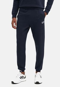 Pantaloni jogger navy con vita elastica e caviglie a coste. Presentano il logo "EA7" sulla coscia sinistra. Realizzati in tessuto morbido e liscio.