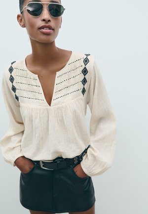 Chemisier crème avec motif brodé noir, décolleté en V et manches bouffantes, associé à une mini-jupe en cuir noir et une ceinture cloutée.