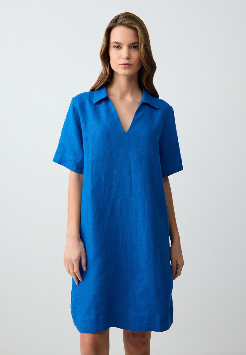 Donna in piedi, che indossa un vestito blu a maniche corte e all'altezza del ginocchio con colletto a V, su uno sfondo grigio chiaro uniforme.