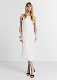 Frau mit langen blonden Haaren, die ein ärmelloses weißes Midikleid und schwarze High-Heel-Sandaletten vor einem schlichten weißen Hintergrund trägt.