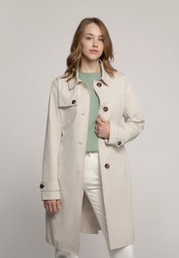 Beige trenchcoat met een kraag, bruine knopen en lange mouwen. Gemaakt over een lichtgroene top en witte broek, met een elegante uitstraling.