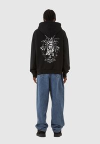 Zwarte hoodie met een achterafbeelding van een figuur omringd door rozen; spijkerbroek met een relaxte pasvorm; zwarte schoenen.