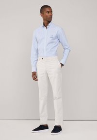 Camisa de manga larga a rayas azul claro con cuello de botones, combinada con pantalones blancos y zapatillas oscuras azules, de pie frente a un fondo neutro.