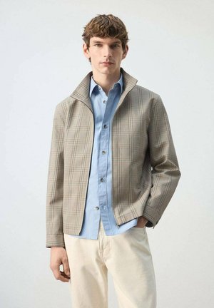 Joven con chaqueta beige de cuadros con cremallera sobre una camisa azul claro abotonada y pantalones color crema, de pie contra un fondo liso.