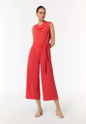 Roter Jumpsuit mit Drapé-Ausschnitt, weit geschnittene Hosen und einem Stoffgürtel. Glatte Textur; kombiniert mit hellen, geschnürten Absätzen.