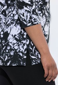 Schwarz-weißes Oberteil mit floralem Muster, abgerundetem Saum, seitlichen Schlitzen, Dreiviertelärmeln und gerippter Textur. Weicher Stoff-Finish.