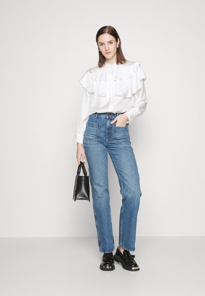 Blusa blanca con volantes en capas, jeans de tiro alto azules y zapatos de charol negros. Sosteniendo un pequeño bolso negro. Fondo simple.