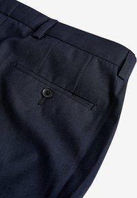 Marineblaue maßgeschneiderte Hosen aus einem strukturierten Stoff, ausgestattet mit einer einzelnen Gesäßtasche und einem schwarzen Knopfverschluss. Verstärkter Bund.