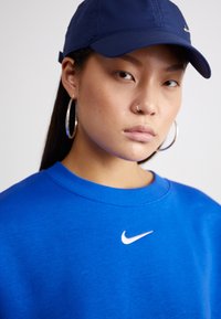 Blå sweatshirt med rundad halsringning och vitt Nike-logotyp; ihopparad med en mörkblå basebollkeps och stora silverfärgade hoop-örhängen.