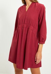 Rotes, leichtes Kleid mit V-Ausschnitt, dreiviertel Ärmeln, geraffter Taille und seitlichen Taschen. Glatte Textur, lässige Passform, knielange Länge.