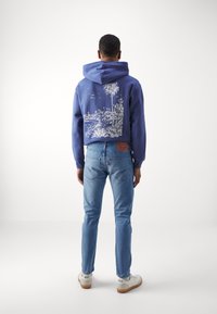 Man som bär en blå hoodie med vit tropisk landskapsprint, ljusblå jeans och vita Nike sneakers, står vänd bortåt mot en enfärgad bakgrund.