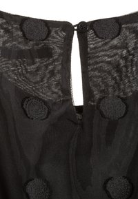 Genomskinlig svart tyg med stora texturerade prickar, med en liten knapp och ögla som stängning i övre mitten.