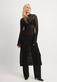 Black knitted dress with a long silhouette, featuring an open mesh pattern, long sleeves, and a high neckline. Worn over black trousers.