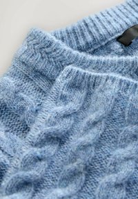 Lyseblå strikket sweater med kabelmønster, ribbet halsudskæring og blød tekstur. Stoffet ser fuzzy og varmt ud.