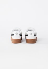 Ein Paar weiße Puma-Sneaker mit braunen Sohlen, schwarzen Seitenstreifen und Puma-Logo auf der Ferse, von hinten gegen einen weißen Hintergrund betrachtet.